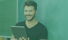 PS Professor(a) Conteudista UAB Homem jovem, branco, magro, de cabelo liso e barba sorrindo enquanto segura um tablet prata. Ele usa uma camiseta preta e ao fundo, vê-se uma lousa verde e uma parede ladrilhada.